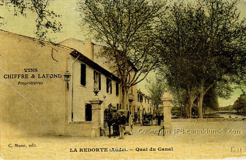 Sur cette carte postale colorisée, on aperçoit les établissements d'un vigneron. Sur le mur de la maison, à gauche, est inscrit : Vins CHIFFRE et LAFOND, propriétaires. À l'entrée, un attelage est stationné et nous fait face, chargé de vins. Un homme coiffé d'un béret tient le cheval par la bride. Plus loin, dans la cour, on aperçoit une voiture ancienne. Le Canal du Midi est également visible, sur la droite.