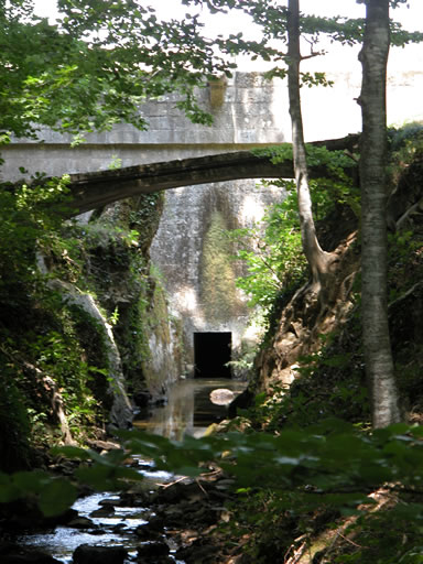 Sous les frondaisons. Un petit pont photographié en contre-plongée. Il franchit le petit canal destiné à évacuer le trop plein du barrage du Lampy. Au fond, vue de la vanne d'évacuation.