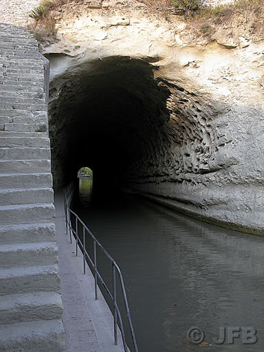 Le tunnel du Malpas vu depuis le côté Ouest. C'est la fin de l'après midi, le soleil vient lécher les parois d'entrée du tunnel. Après l'ombre, on aperçoit la sortie. A gauche, un escalier permet de rejoindre le sommet (la prise de vue est verticale)