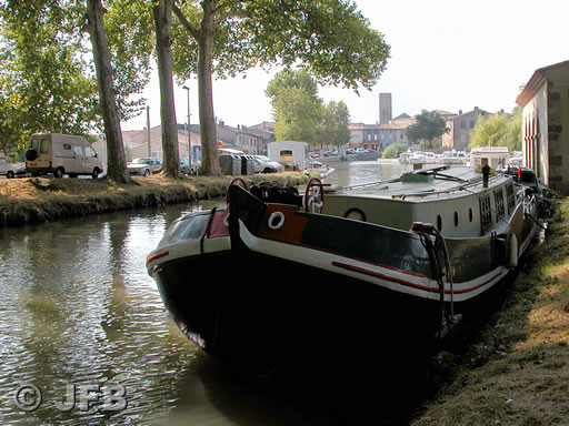 Il s'agit de la péniche verte et blanche, soulignée de rouge, déjà décrite sur une autre photo. Ici, la vue est de proue, dans l'ombre des platanes du Canal du Midi et en arrière plan, on aperçoit la petite ville de Trèbes noyée dans le soleil de l'été.