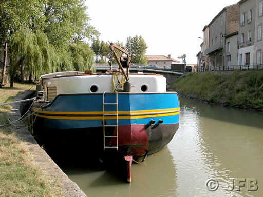 Il s'agit de la péniche bleue et noire, déjà décrite sur une autre photo. Ici, la vue est de proue. On aperçoit très bien le gouvernail. Une échelle est accroché, certainement pour permettre quelques travaux sur le gouvernail. De l'autre côté du Canal du Midi, on aperçoit les maisons de Trèbes.