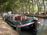 Péniche originale amarrée près de Trèbes. Elle est de couleur verte, avec une ligne blanche et épaisse complétée par des lignes rouges, plus fines. La cabine est blanche, agrémentée par de belles boiseries. Comme la vue est de proue, on aperçoit le timon, qui est énorme, taillé dans le bois, verni et sculpté à l'ancienne.