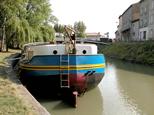 Il s'agit de la péniche bleue et noire, déjà décrite sur une autre photo. Ici, la vue est de proue. On aperçoit très bien le gouvernail. Une échelle est accroché, certainement pour permettre quelques travaux sur le gouvernail. De l'autre côté du Canal du Midi, on aperçoit les maisons de Trèbes.