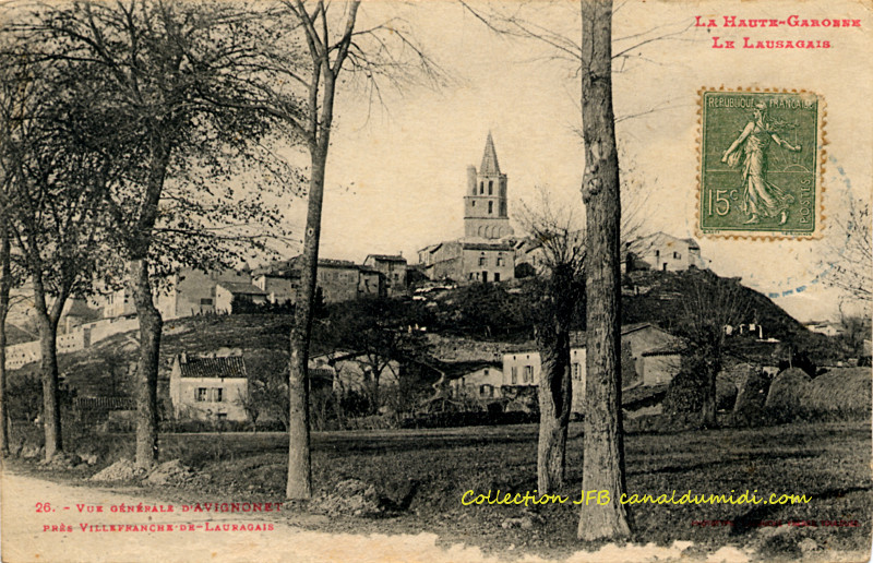 Carte postale ancienne, légendée : La Haute-Garonne - Le Lauragais - Vue générale d'Avignonet près Villefranche de Lauragais. On aperçoit une partie de la route, et quatre arbres qui la bordent. Plus loin dans un pré, un mûrier. Le c&œlig;ur du village est situé sur une petite colline et l'église domine. Au pied de la colline, quelques maisons.