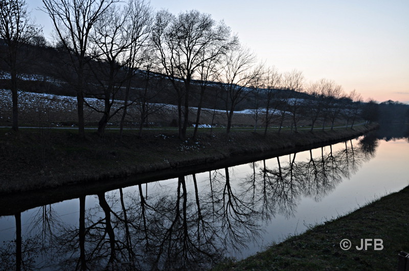 Le canal du Midi au crépuscule, près de l'écluse d'Emborrel : On aperçoit une rangée de jeunes platanes 
qui se reflètent dans l'eau du canal. Sur les francs bords et sur les pentes des prairies avoisinantes, on aperçoit u
n peu de neige et à l'horizon, les dernières lueurs du soleil déjà couché.