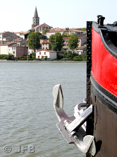 L'avant d'une belle péniche rouge, sur le Grand bassin de Castelnaudary, avec une belle ancre peinte en neuf, et, en toile de fond, l'église et la ville