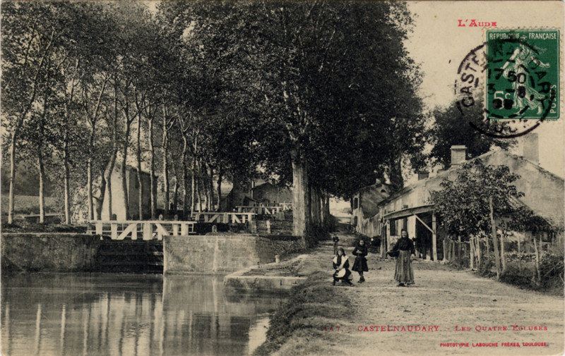 Carte postale ancienne légendée : Castelnaudary, les quatre écluses. C'est une vue d'amont. On aperçoit bien les quatre vantaux dont l'armature est en bois. À gauche du canal, on voit le moulin, et à droite, quelques maisons, une femme portant un panier et deux enfants. Plus loin, toujours à droite, un homme vient vers nous. Le canal est bordé de jeunes platanes.