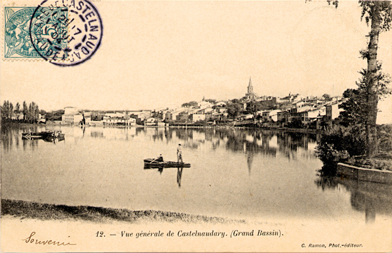 Carte postale ancienne légendée : Vue générale de Castelnaudary(grand bassin). On aperçoit le grand bassin en premier plan. De l'autre côté
    de la rive, la ville de Castelnaudary avec l'église qui domine. Au milieu du bassin, des embarcations stationnent, notamment une barque sur laquelle un homme tient les rames et un autre est debout.