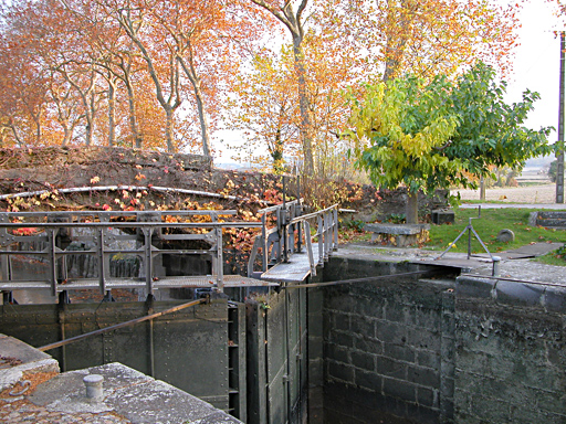 Gros plan sur la vanne de l'écluse, en arrière plan, un petit pont et quelques arbres aux couleurs automnales variées