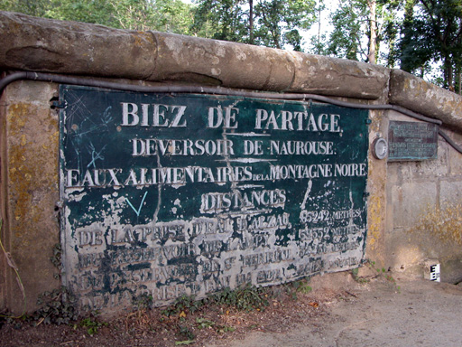 Plaque d'identification du bief de partage sur laquelle est écrit (orthographe d'époque) : BIEZ DE PARTAGE - DEVERSOIR DE NAUROUSE - EAUX ALIMENTAIRES DE LA MONTAGNE NOIRE - DISTANCES - DE LA PRISE D'EAU 65242 mètres - DU RESERVOIR DU LAMPY 52552 mètres - DU RESERVOIR DE SAINT-FERREOL 34456 mètres