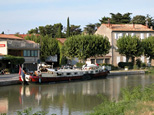 Le petit port de La Redorte : On aperçoit les bâtiments du port, quelques arbres (nous sommes en été) et une fastueuse péniche amarrée... Sa coque est de couleur rouge pour la partie supérieure, et noire, pour la partie inférieure. Les structures du pont sont d'un blanc immaculé. On distingue 7 fenêtres rectangulaires agrémentées de rideaux. La proue, ainsi que les yeux de la péniche, sont peints en blanc. On aperçoit deux drapeaux sur la proue : un rouge, et un bleu. Magnifique !!!