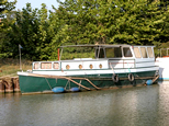 Une péniche faite pour l'habitation, mais quelque peu modeste cependant. La péniche nous fait façe à gauche sur la rive opposée du canal. La coque est entièrement verte. La cabine et le poste de pilotage sont blancs. La partie habitable montre quatre petits hublots ronds sur le côté gauche que nous pouvons apercevoir. Sur la proue, trois hublots rectangulaires, dont le central est plus large. Encore sur la proue, on distingue un petit mat agrémenté d'un fanion bleu.