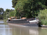 Péniche à dominante gris foncé et une ligne blanche sur la proue, sur laquelle on aperçoit une enseigne, caractères blancs sur fond brun : Menuiserie fluviale. Une autre caractéristique consiste en un rateau télévision dont le mat est considérablement haut