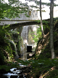 Sous les frondaisons. Un petit pont photographié en contre-plongée. Il franchit le petit canal destiné à évacuer le trop plein du barrage du Lampy. Au fond, vue de la vanne d'évacuation.