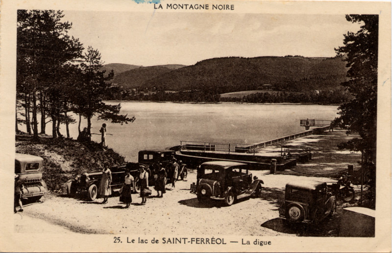 Carte postale ancienne légendée : 
La Montagne Noire - Le Lac de Saint-Ferréol
    - La Digue.  On aperçoit le lac de Saint-Ferréol à son
    niveau presque optimal, le début de la digue et le système d'évacuation
    du trop plein. Quelques tacots des années 30 sont stationnés en premier
    plan, et l'on voit quelques visiteurs vétus à la mode de l'époque