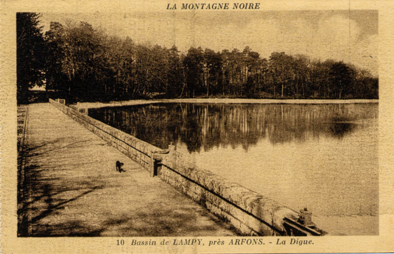 Carte postale ancienne ou l'on voit à gauche, la digue du bassin du Lampy sur lequel se trouve un chien noir. A Gauche, le lac, et au fond, la forêt;