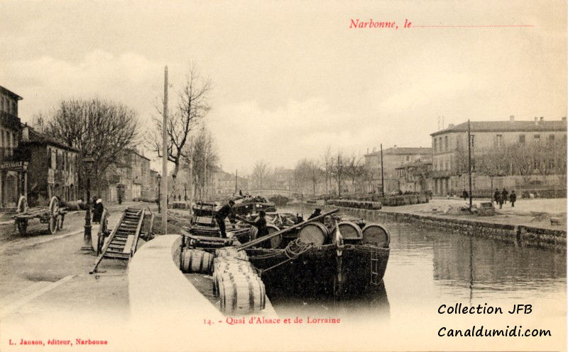 Carte postale ancienne légendée : Narbonne, quai d'Alsace et de Lorraine. On aperçoit une péniche amarrée entièrement chargée de barriques. Les arbres qui bordent le canal sont dénudés, c'est l'hiver. Un homme sur le quai, tend la main à une femme, qui se trouve sur la péniche, probablement pour l'aider à débarquer. Près de là, une charette vide est stationnée. Sur la route qui borde le canal, on aperçoit un attelage.