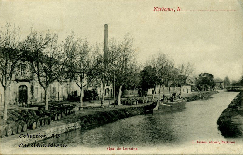 Carte postale ancienne légendée : Narbonne, quai de Lorraine. On aperçoit une péniche amarrée dotée d'un mat. Elle est probablement vide car elle est haute sur l'eau. Trois personnes s'affairent sur le pont. Sur les quais, de nombreuses barriques sont disposées, attendant peut être d'être chargées. D'après l'aspect des arbres, il semble que ce soit le printemps. On aperçoit également en arrière plan, une cheminée d'usine.