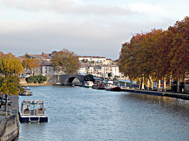 Perspective du port de Castelnaudary depuis le pont neuf par une belle journée d'automne. L'eau est bleue, quelques péniches sont amarrées sur les rives et des platanes bordent les quais. On aperçoit plus loin le pont en dos d'âne qui donne accès au grand bassin