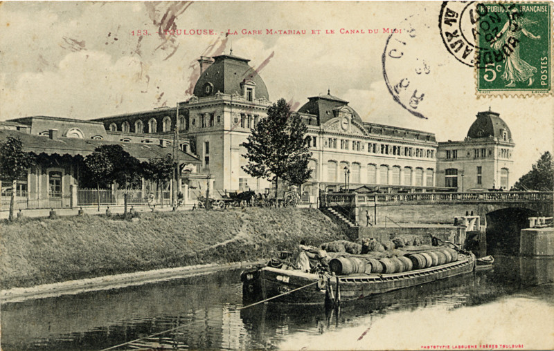 Carte postale ancienne légendée : Toulouse, la gare Matabiau et le Canal du Midi. Cette carte est un peu salie par l'encre, mais elle me plait. On aperçoit au premier plan le canal, avec une péniche chargée de barriques de vin dont le niveau est bas sur l'eau. On voit très bien le cable de halage, mais pas les chevaux qui constituent le moteur. Ensuite, au second plan, on voit très bien la gare Matabiau, avec son horloge...