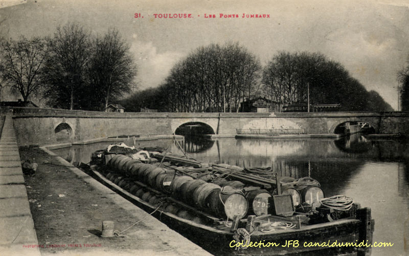 Carte postale ancienne légendée : Toulouse - Les ponts jumeaux. Trois ponts sont représentés : À gauche, c'est le point de départ du canal latéral à la Garonne, au centre, nous avons le Canal du Midi et à droite, séparé par le bas relief, le Canal de Brienne. Au tout premier plan, une péniche chargée de barriques de vin est amarrée.