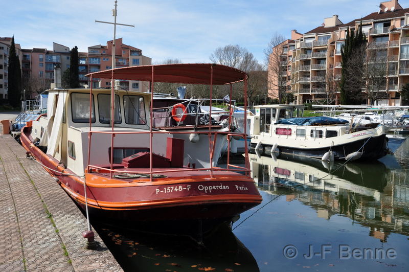 À gauche, une petite péniche de couleurs bordeaux et orange, est amarrée sur les quais de Port Sud. On aperçoit une partie du bassin, et au second plan, d'autres bateaux qui se reflètent dans l'eau bleue. Plus loin, les résidences modernes qui entourent le bassin.