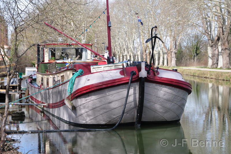 Péniche d'habitation grise avec des lignes rouges d'un très bel effet. Elle dispose d'un mat également peint en rouge. La péniche est amarrée sur le Canal du Midi.