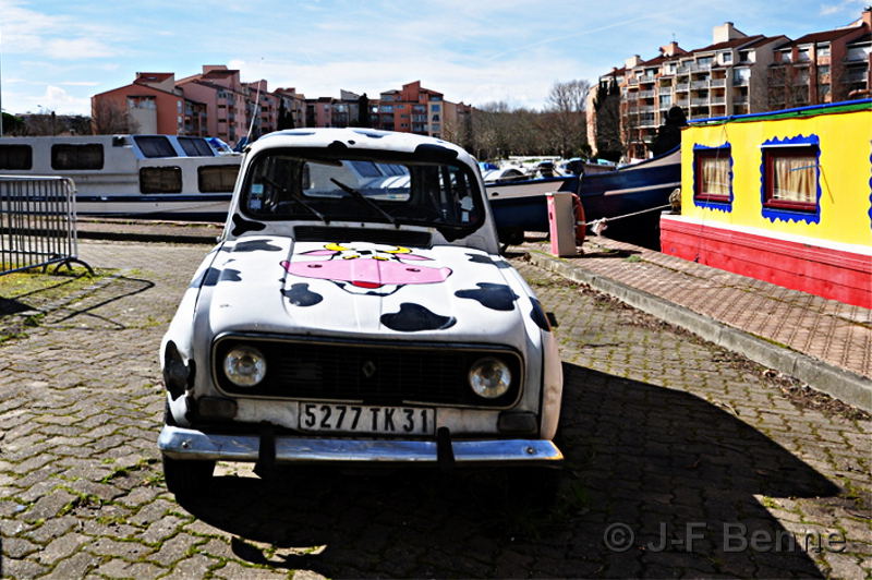 Une voiture 4L décorée humoristiquement en blanc et noir à la manière d'une vache normande, et pour rigoler encore plus, sur le capot, le beau portrait de face d'une vache non moins rigolote. Le véhicule est photographié de face. À droite de la voiture, une péniche étrange peinte en jaune vif.